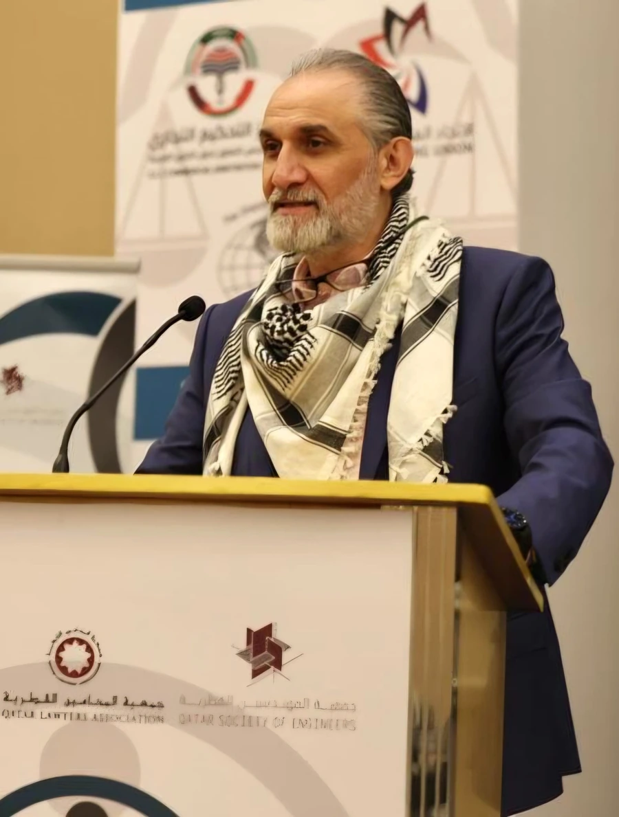 Ammar Anabtawi speaking at a podium in a blue suit and keffiyeh scarf